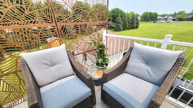 Two wicker chairs with grey cushions on a patterned floor, a decorative metal fence, and a view of a green lawn and parked cars.
