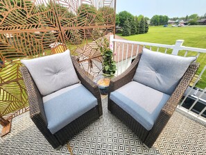 Twee rieten stoelen met grijze kussens op een patroon vloer, een decoratieve metalen hek en uitzicht op een groen gazon en geparkeerde auto's.