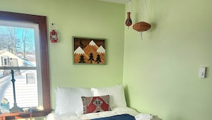 A neatly made bed with a colorful blanket, a bedside table with a lamp, and a painting of mountains on the wall.