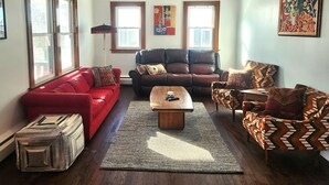 A living room with a red sofa, a brown leather couch, and patterned armchairs. There is a wooden coffee table in the center, a ceiling fan, and artwork on the walls.