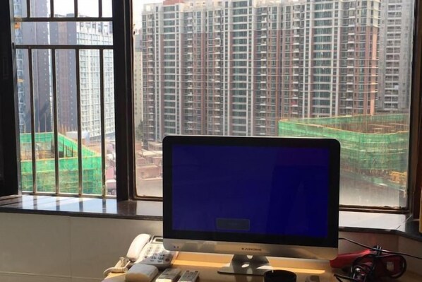 Une pièce avec un écran d’ordinateur, un téléphone et une paire de ciseaux sur un bureau. Une fenêtre donne sur des immeubles de grande hauteur et un chantier de construction.