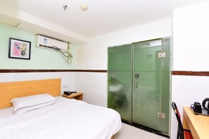 A hotel room with a bed, a green wardrobe, a small bedside table with a lamp, and a wall-mounted air conditioner.