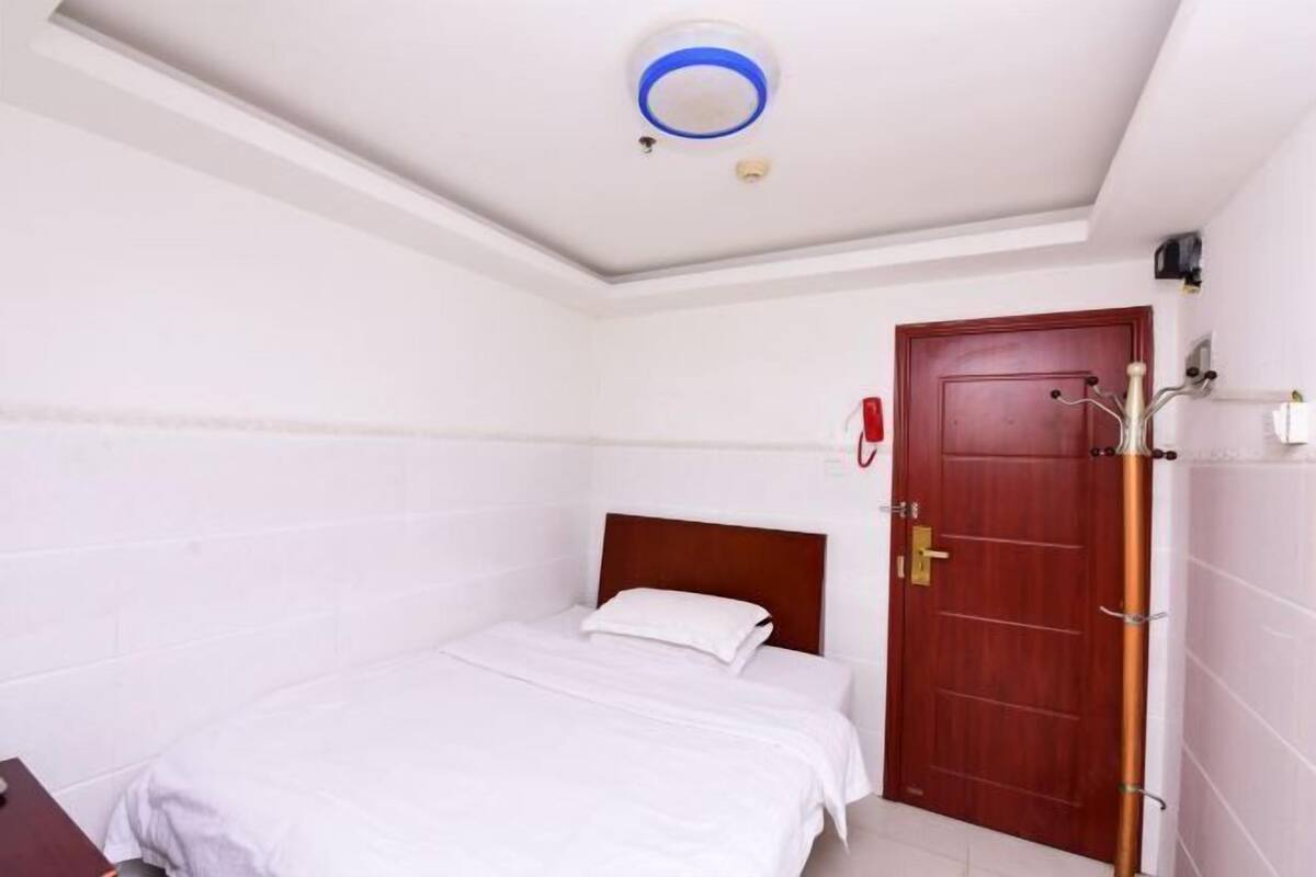 Una cama individual con ropa blanca, cabecera de madera, una puerta roja y un teléfono fijo en la pared.