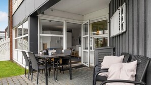 Un patio exterior con una mesa y sillas, vista hacia el interior de la casa y una pared exterior gris.