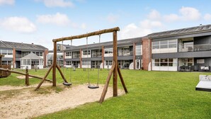 Un parque infantil con columpios y un arenero, rodeado por un edificio con varias ventanas y balcones.