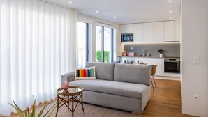 A modern living room with a grey sofa, a small round table, and a colorful pillow. A kitchen is visible in the background with white cabinets and stainless steel appliances. - Pestana Apartments T1 Terrace  (Porto)