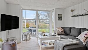 Uma sala de estar moderna com uma televisão grande, uma mesa de centro de vidro, um sofá cinza e vista para o exterior através de uma porta de correr de vidro.