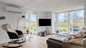 Sala moderna com televisão de tela plana na parede, uma poltrona de balanço, uma mesa de centro com objetos decorativos e uma grande janela com vista para o exterior.