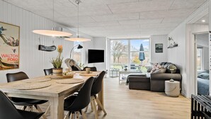 Uma sala ampla com mesa de jantar de madeira, cadeiras pretas, sofá cinza e uma grande janela com vista para o exterior.