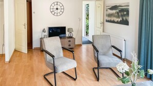 Un salon avec deux fauteuils gris rembourrés, un téléviseur, une horloge et un tableau représentant une forêt.