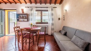 A cozy living area with a dining table, chairs, a sofa, a window with curtains, a radiator, and a shelf with decorative items.