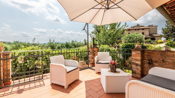 A terrace with wicker furniture, a large umbrella, and a view of greenery and buildings.