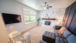 A living room with a flat-screen TV, a stone wall, a fireplace, and a ceiling fan. - Charming Historic Home with chef's kitchen formal dining, sitting room, & patio (Payson)
