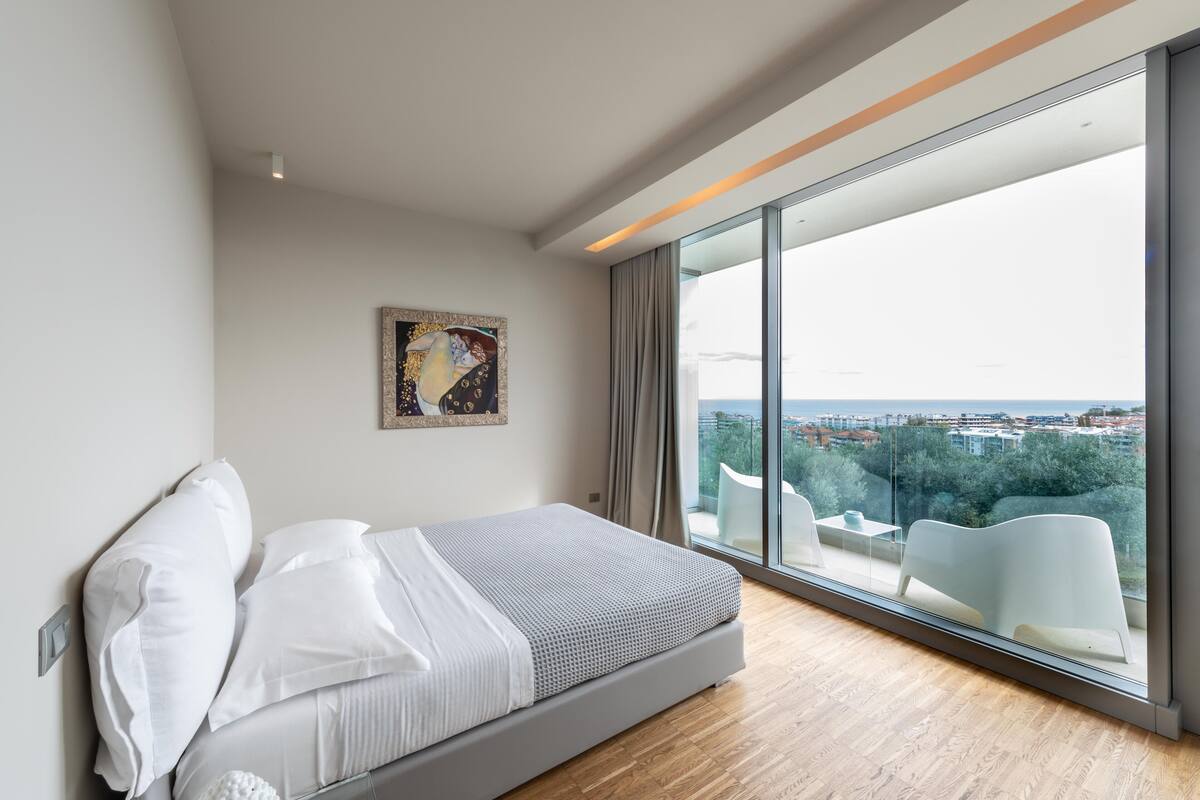 Une chambre moderne dotée d’un grand lit, offrant une vue sur la ville grâce à de larges fenêtres du sol au plafond, et présentant un design minimaliste.