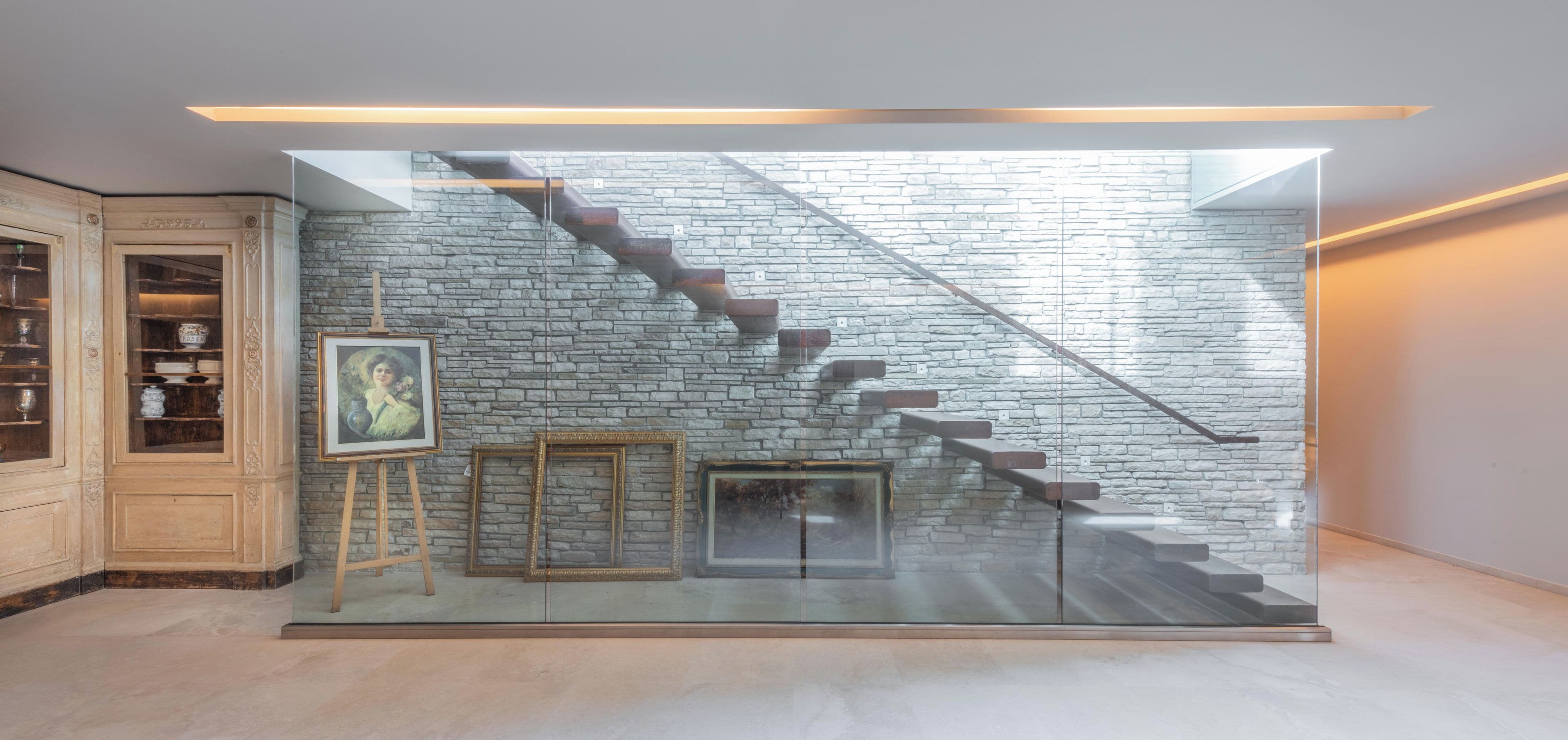 A modern interior with a glass staircase against a stone wall, wooden cabinets, and framed artwork.