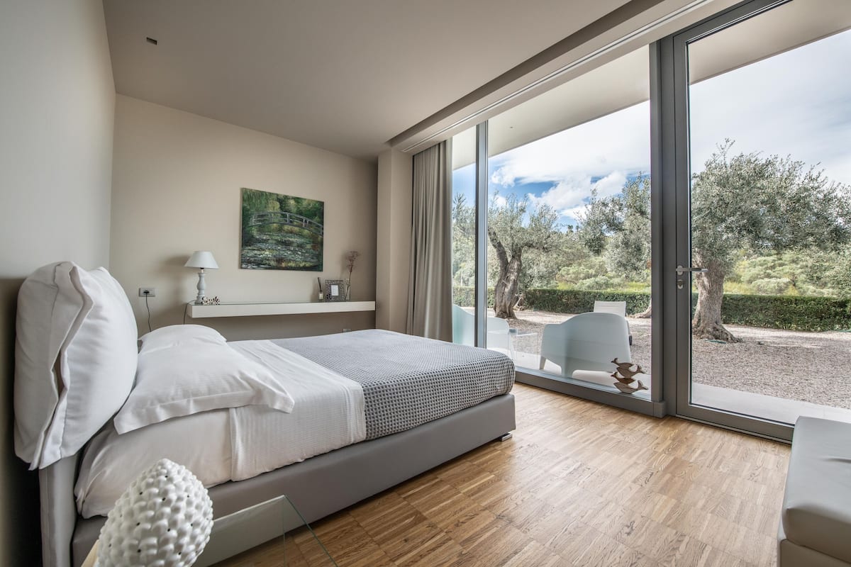 Une chambre à coucher moderne avec un grand lit, une table de chevet avec une lampe, un tableau et une vue sur l’extérieur.