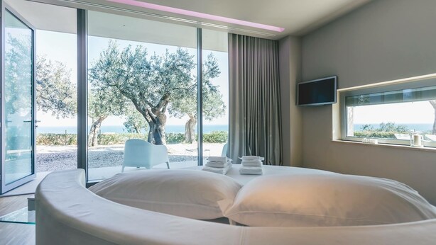 Une chambre moderne avec un lit aux lignes courbes, un téléviseur fixé au mur et une vue sur la mer et les arbres, grâce à de grandes portes vitrées.