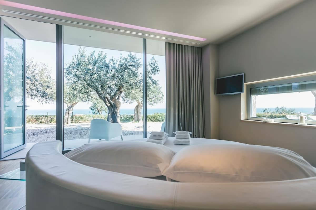 Une chambre moderne avec un lit aux lignes courbes, un téléviseur fixé au mur et une vue sur la mer et les arbres, grâce à de grandes portes vitrées.