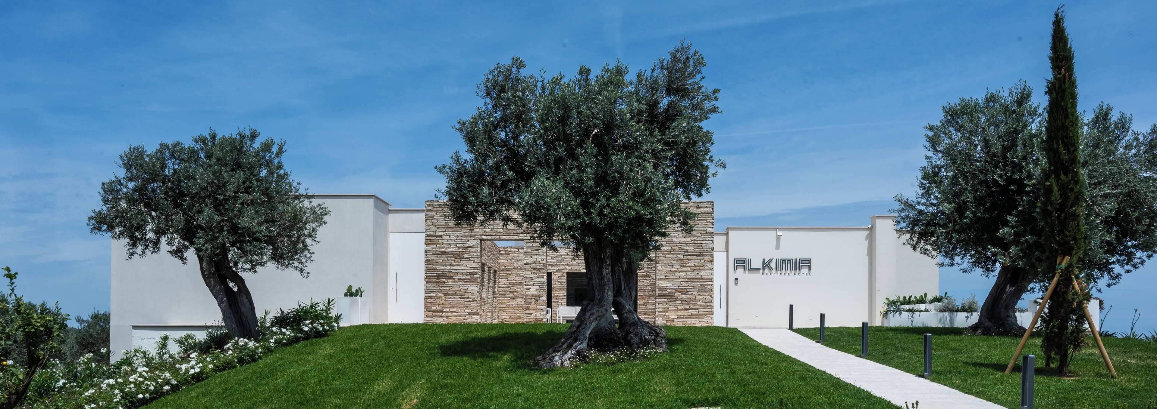 A modern hotel with a stone facade, named 'ALKIMIA BOUTIQUE HOTEL,' surrounded by well-maintained greenery and a clear sky.