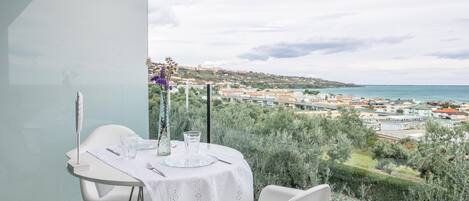A dining table set with a white tablecloth, a vase with purple flowers, and clear glasses. The table is positioned to overlook a scenic view of a coastal town and the sea.