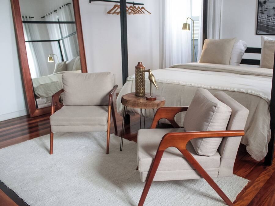 Une chambre d’hôtel moderne comprenant un lit, deux fauteuils, une petite table ronde ornée d’une sculpture d’oiseau, un miroir et une fenêtre avec des rideaux.