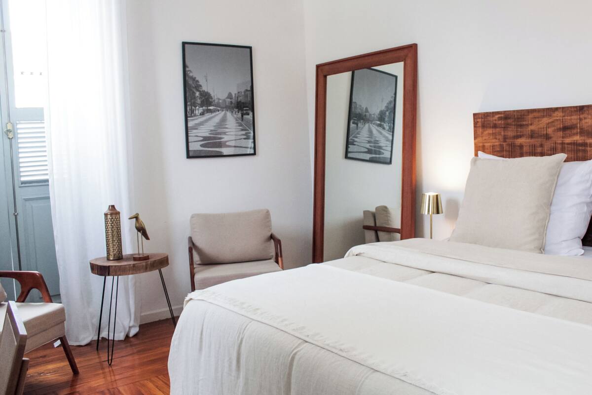 A hotel room with a bed, a chair, a small table, a mirror, and two framed pictures on the wall.