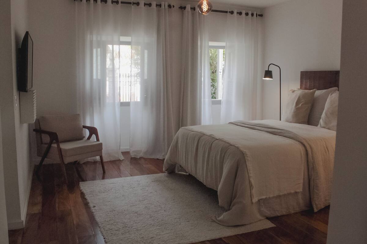A bedroom with a bed, a chair, a rug, a TV, and a window with curtains.
