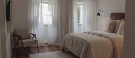 Ein Schlafzimmer mit einem Bett, einem Sessel, einem Teppich, einem Fernseher und einem Fenster mit Vorhängen.
