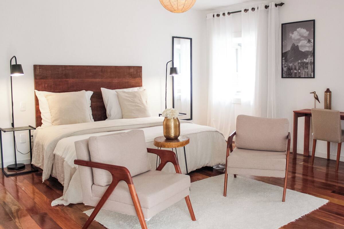 A modern bedroom with a wooden headboard, a large bed, two chairs, a small table, and a hanging light fixture.