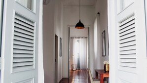 A hallway with a wooden floor, white walls, and a ceiling with a hanging light fixture. There are doors with shutters on either side, and a table with a vase on the right. - Boutique Hotel Castelinho (Rio de Janeiro)
