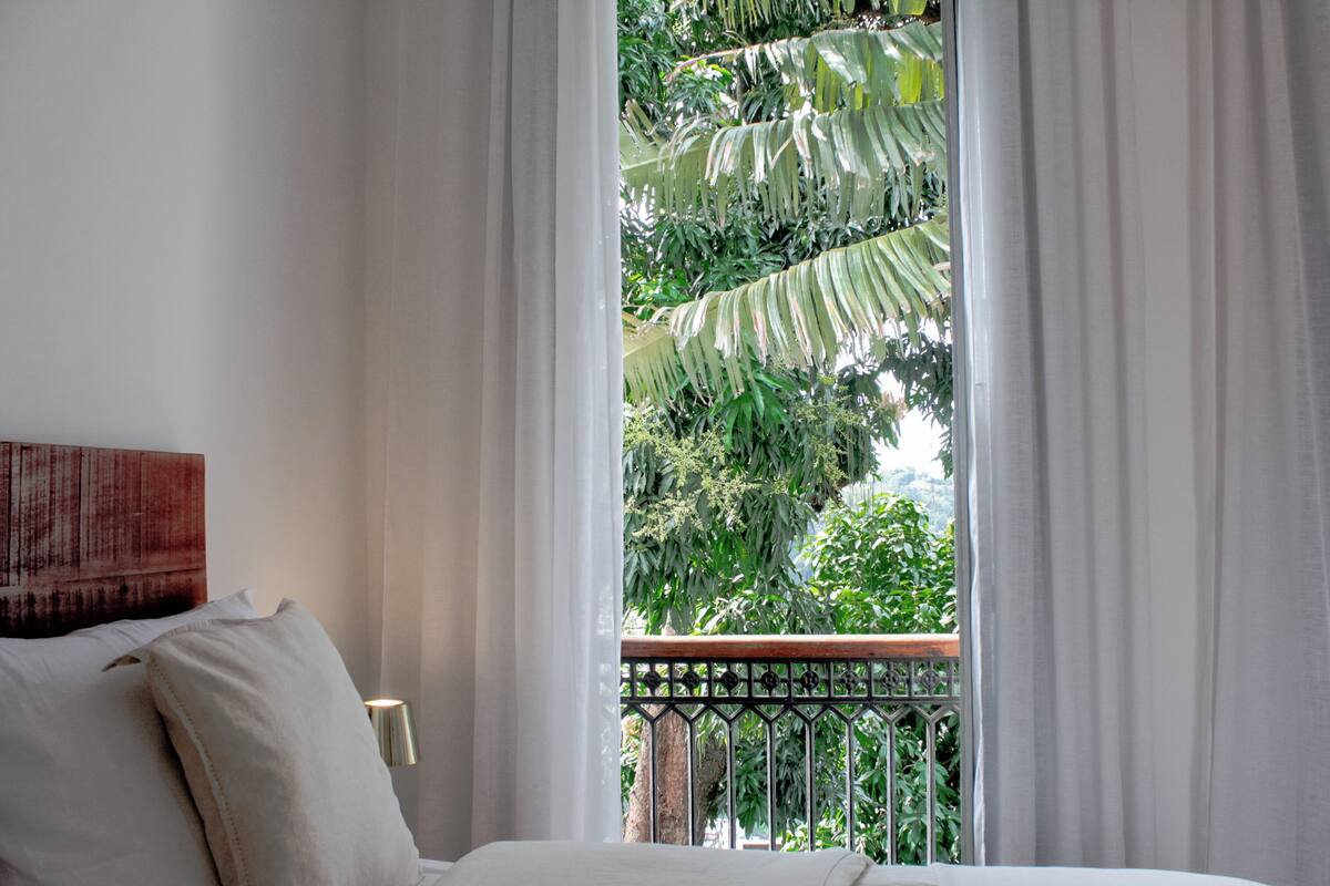 A bedroom with a bed, white curtains, a view of greenery outside, and a metal railing.