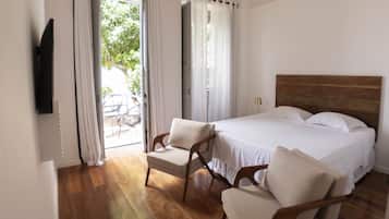 Un dormitorio con suelo de madera, una cama con sábanas blancas, dos sillones blancos y una lámpara en el techo.