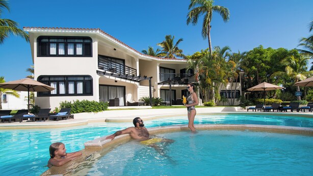 A pool area with lounge chairs and umbrellas, a two-story building with balconies, and palm trees. - Luxury Apartment-Mandatory All-Inclusive (Puerto Plata)