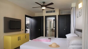 A hotel room with a bed, a television, a ceiling fan, a yellow cabinet, and a bathroom visible through a glass door. - Luxury Apartment-Mandatory All-Inclusive (Puerto Plata)
