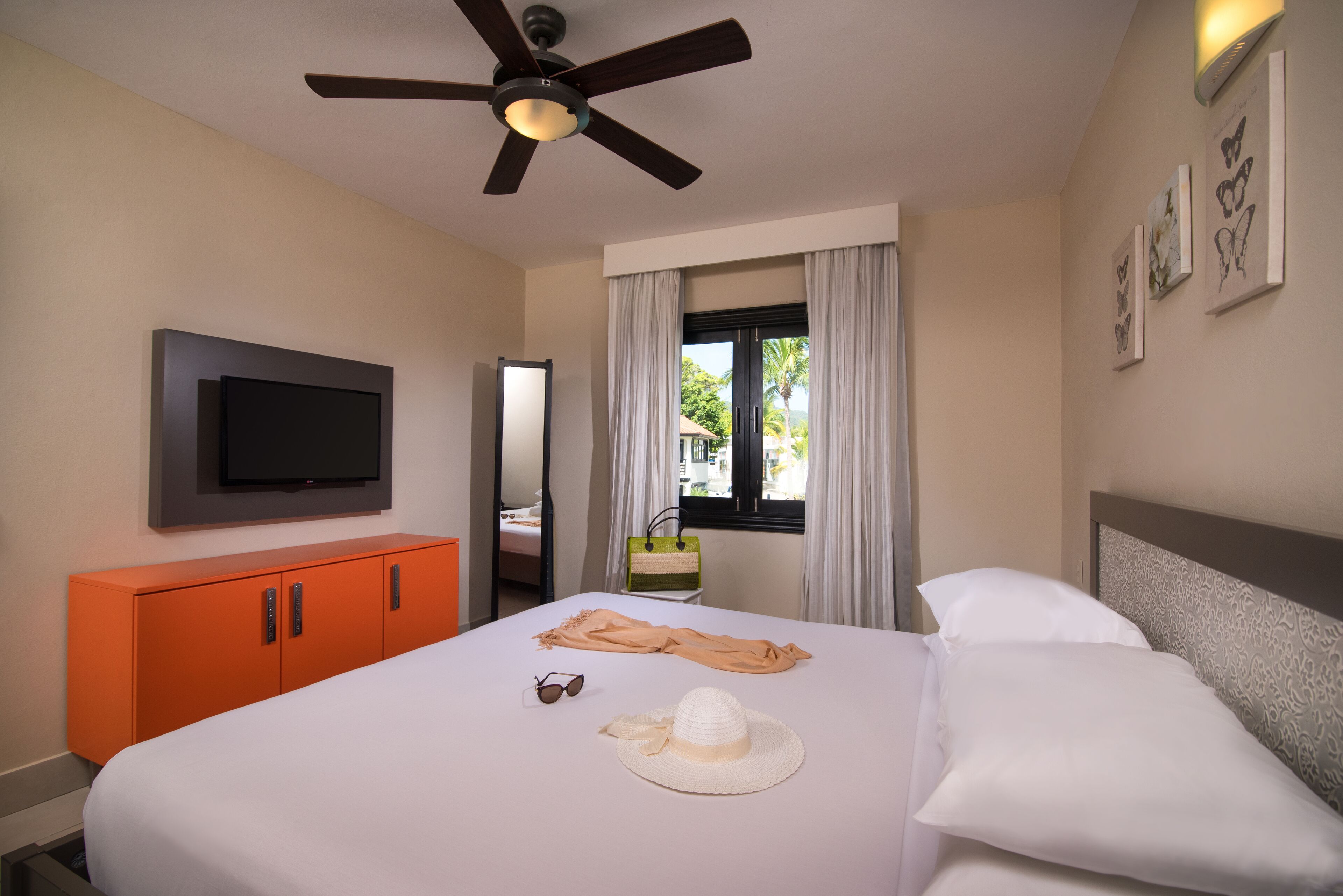 Une chambre d’hôtel avec un lit, une télévision, une armoire orange, un ventilateur de plafond, une fenêtre avec des rideaux et des œuvres d’art accrochées au mur.