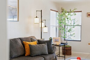 Una sala moderna con un sofá gris, cojines estampados, una mesita con un florero y un ventanal con vistas a zonas verdes.
