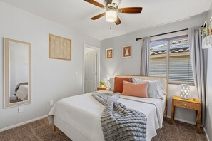 Un dormitorio con cama, ventilador de techo, ventana con cortinas y mesita de noche con lámpara.