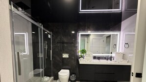 A modern bathroom with a glass shower enclosure, a white toilet, a large mirror, and a vanity with a sink and towel rack. - The Summit 1305 (Panama City Beach)