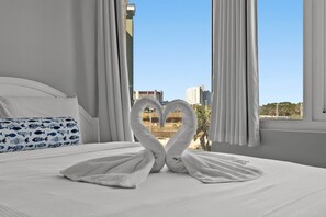 A hotel room with a bed, white curtains, and a view of buildings and palm trees through the window. - Top of The Gulf 306 (Panama City Beach)