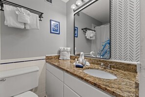 A modern bathroom with a granite countertop, white sink, and a large mirror. There are towels hanging on a rack and a framed picture on the wall. - Top of The Gulf 306 (Panama City Beach)