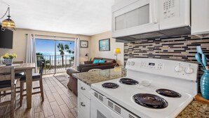 A kitchen with a stove, microwave, and cabinets. There is a dining table with chairs and a view of the ocean through large windows.