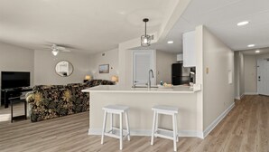 A modern kitchen with a central island, white bar stools, a ceiling fan, and a flat-screen TV in a living area with a floral sofa.