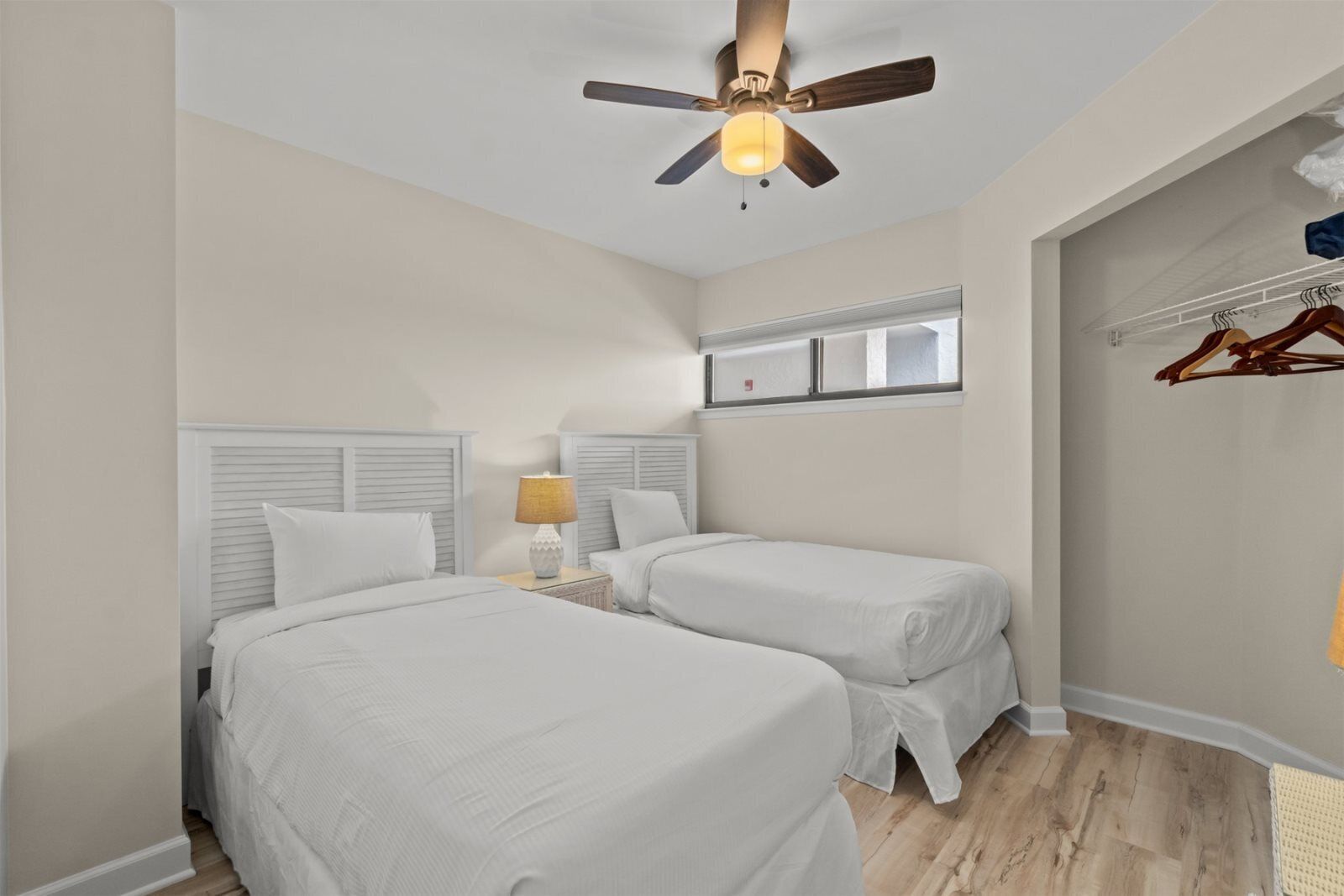 A bedroom with two beds, a ceiling fan, a nightstand with a lamp, a window with blinds, and a coat rack with hangers.