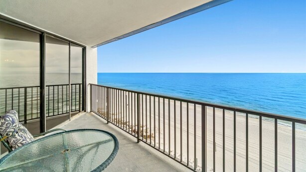 A balcony with a glass table and chairs, overlooking a beach and the ocean.