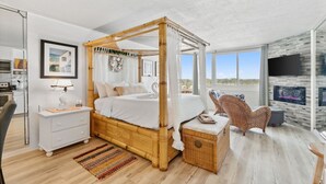 Un dormitorio moderno con cama con dosel, una mesita de noche blanca, una mesita auxiliar de mimbre y vistas al exterior a través de amplios ventanales.