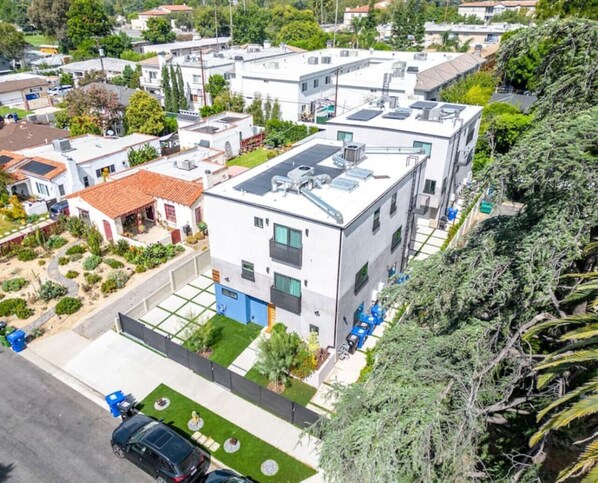 A modern multi-story residential building with a flat roof, solar panels, and a small garden area.