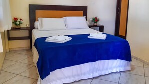 Una cama bien tendida con ropa de cama blanca y azul, dos mesitas de noche con lámparas, y una puerta a la derecha.