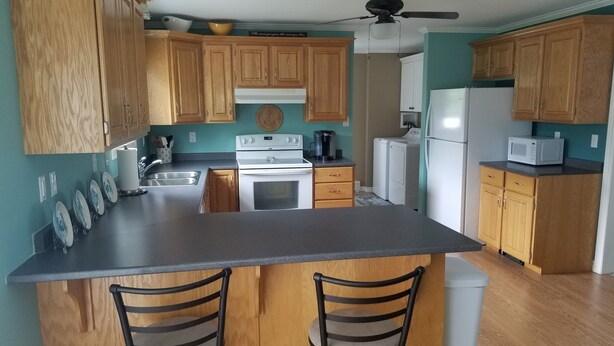 A kitchen with wooden cabinets, a central island with two bar stools, a ceiling fan, and a microwave. - Riverview Home in Milbridge Maine (Milbridge)