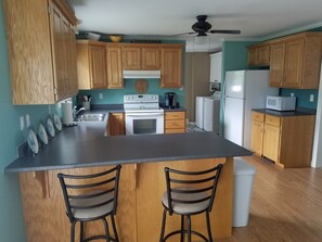 Una cocina con armarios de madera, una isla central con dos taburetes de bar, un ventilador de techo y un microondas.