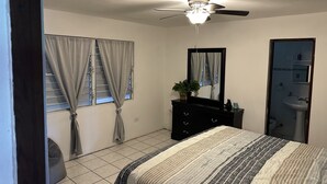 Quarto com cama, ventoinha no teto, cômoda, espelho, janela com cortinas e banheiro com pia e espelho.